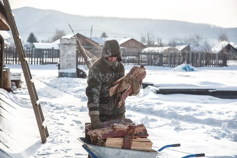 A man in winter clothing handles firewood in a snowy village landscape.