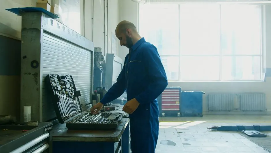 Mechanic inspecting tools in a workshop, wearing a blue jumpsuit with bright natural light from windows.